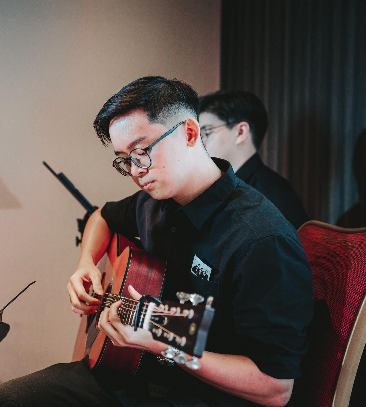 Adriel Tang playing acoustic guitar with a capo on the fretboard, seated in a black shirt during a live performance, with another musician visible behind him.